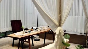 Un comedor con una mesa de madera dispuesta para cuatro personas, un florero con una planta y una ventana con cortinas de encaje.