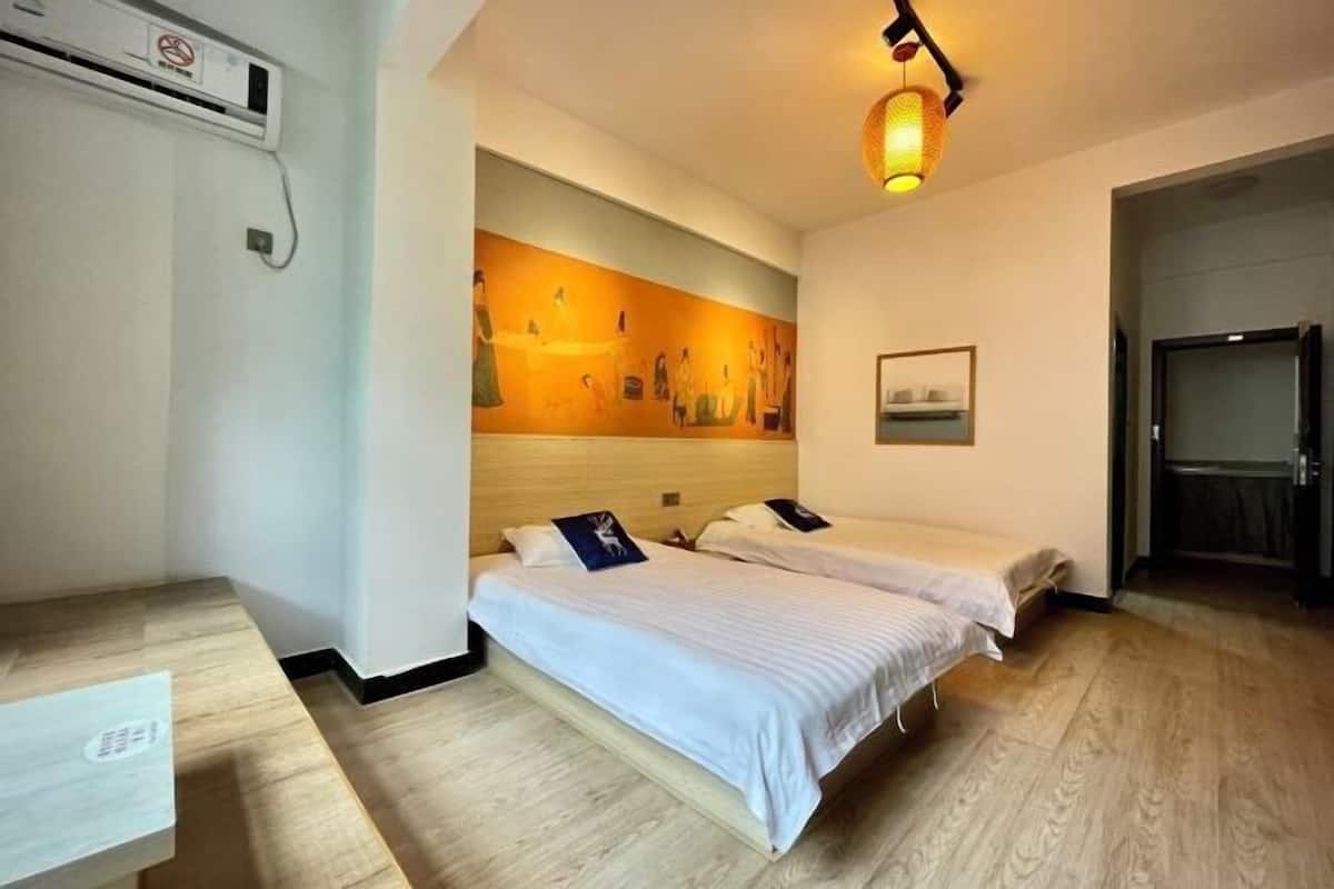 A hotel room with two beds, a wooden floor, a wall-mounted air conditioner, and a painting.
