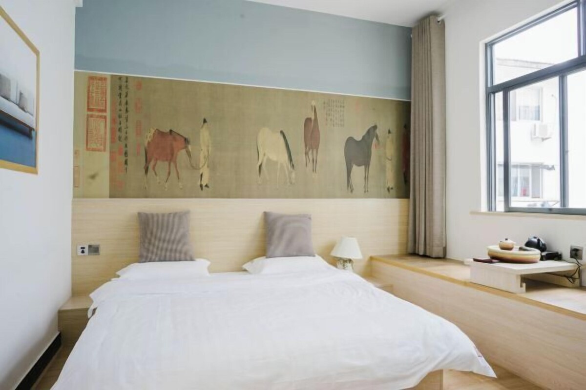 A hotel room with a large bed, a headboard with artwork, a window with curtains, and a bedside table with a lamp and a small decorative item.