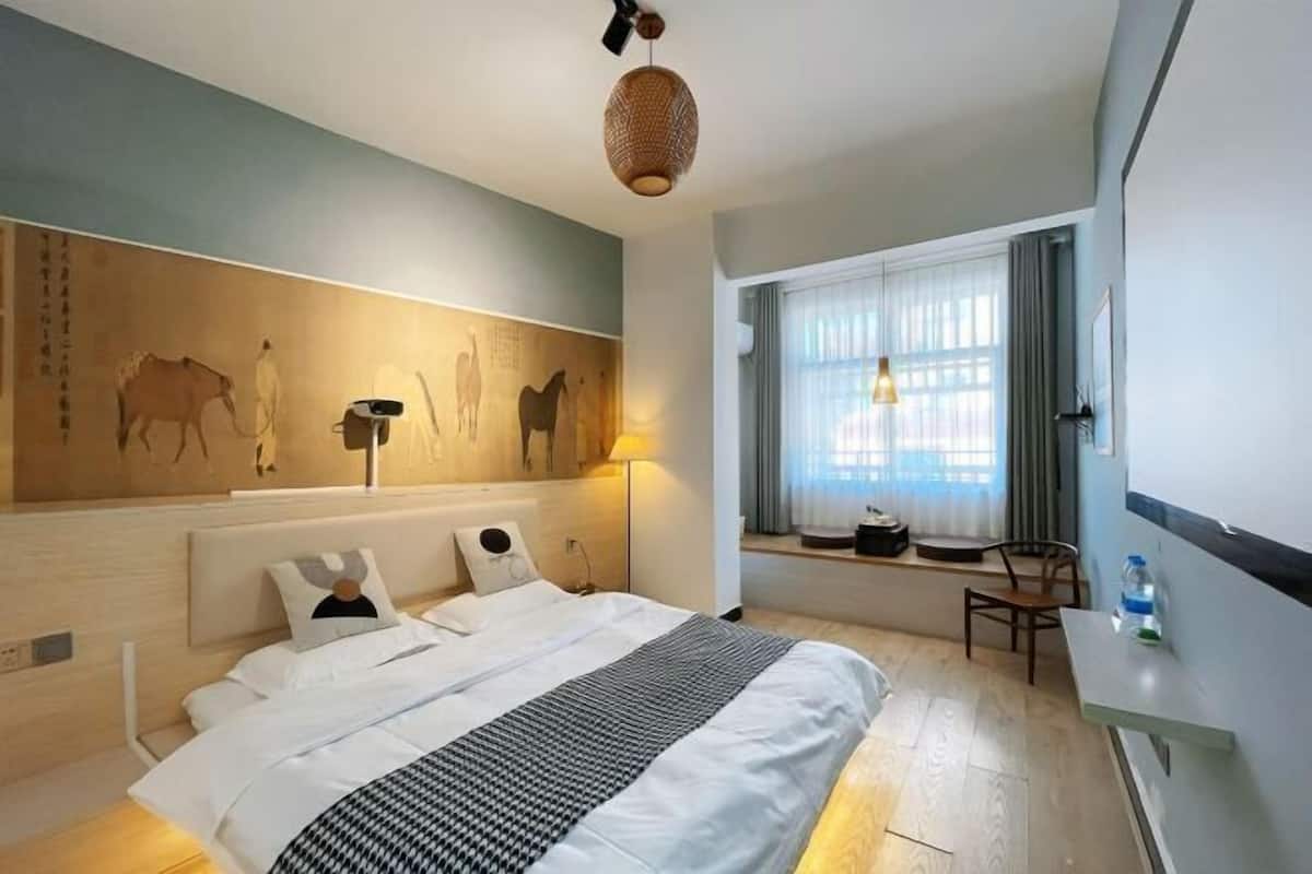 A hotel room with a bed, a desk, a chair, a window with curtains, and a wall with artwork.