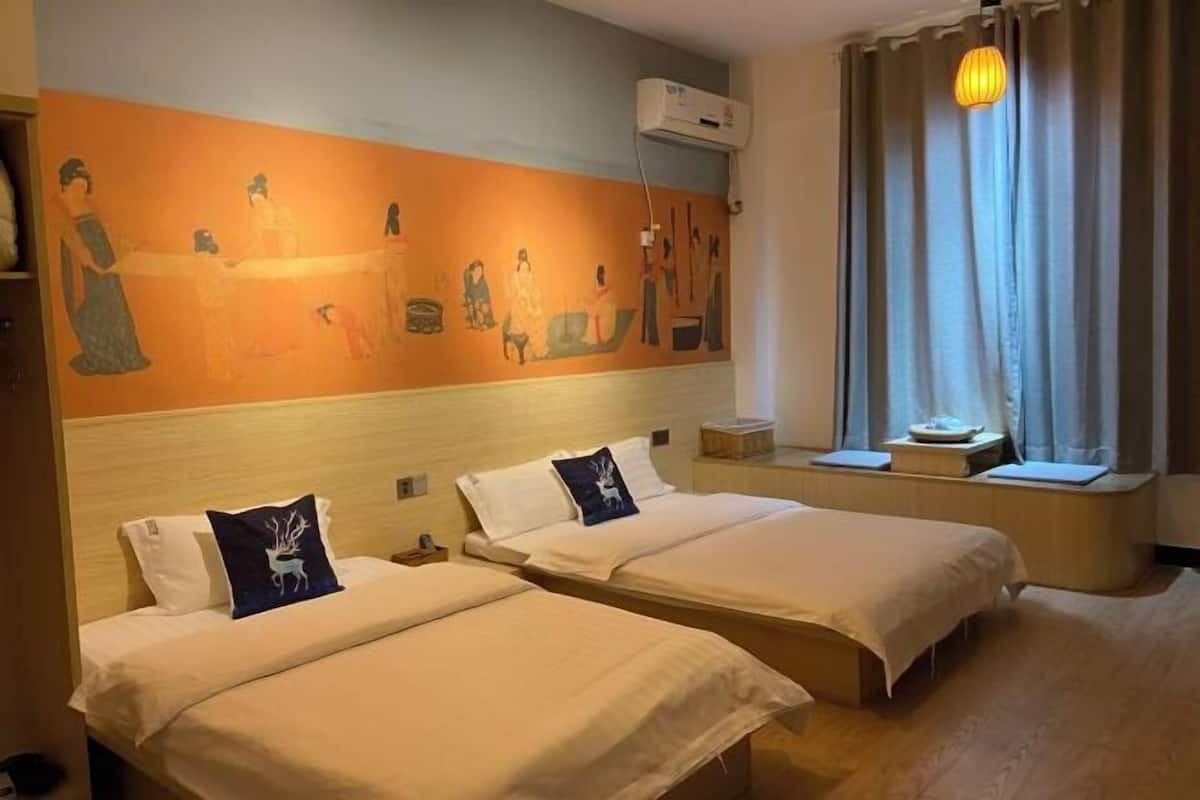 A hotel room with two beds, a mural on the wall, an air conditioning unit, a window with curtains, and a bedside table with a lamp.