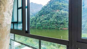 Quarto com uma janela ampla com vista para um rio e uma colina arborizada.