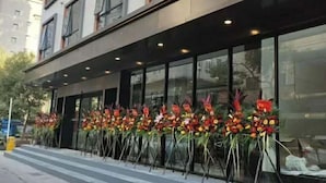 Fachada de edificio con amplios ventanales y una fila de arreglos florales en el exterior.