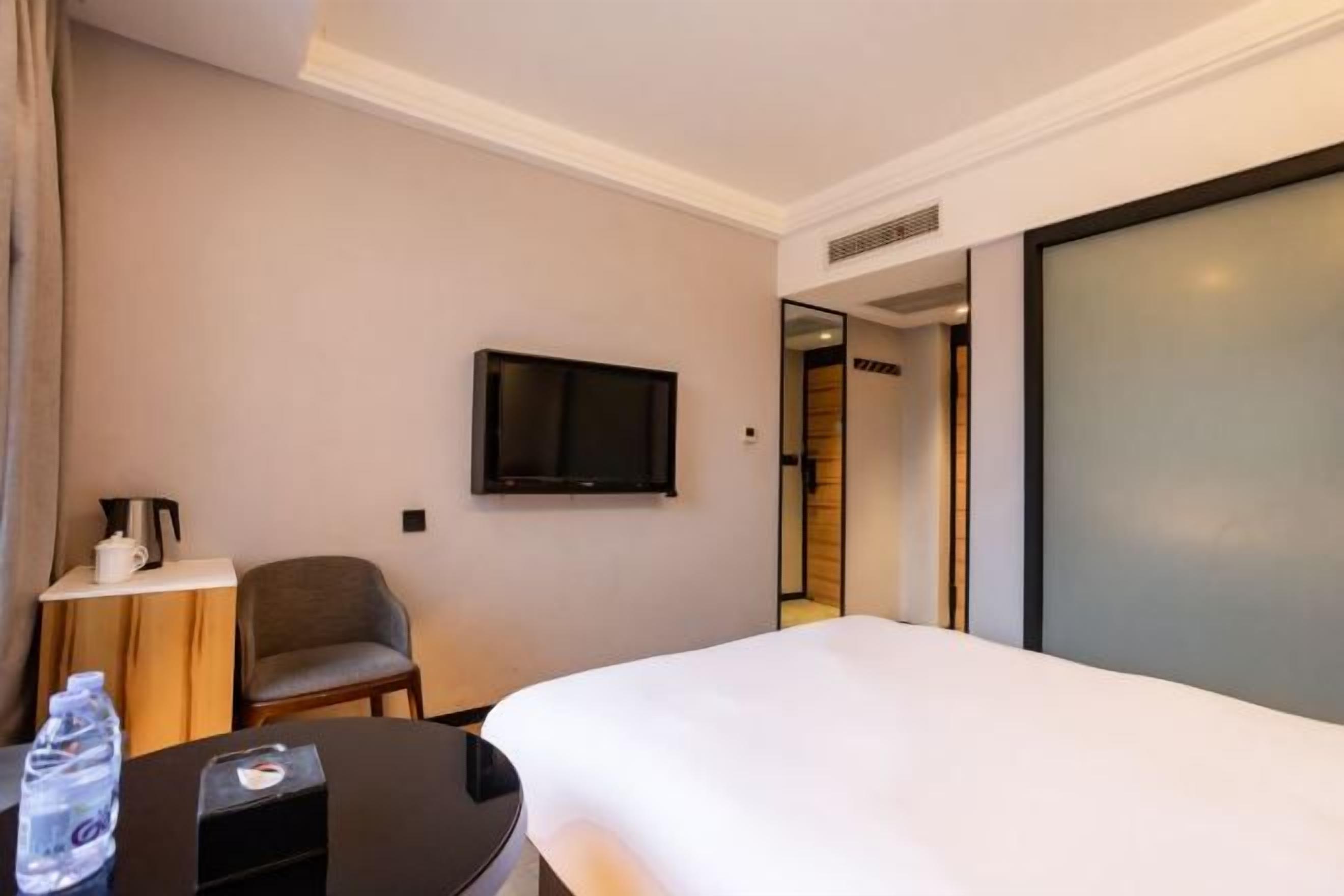 A hotel room with a bed, a television mounted on the wall, a chair, a small table with a water bottle, and a door leading to another room.
