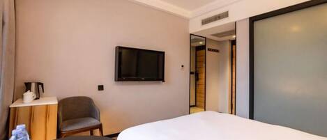 A hotel room with a bed, a television mounted on the wall, a chair, a small table with a water bottle, and a door leading to another room.