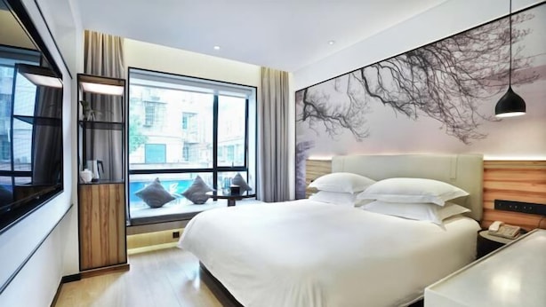 Una habitación de hotel moderna con una cama grande, un ventanal con vistas a edificios, una obra de arte colgada en la pared y una mesita de noche con lámpara.