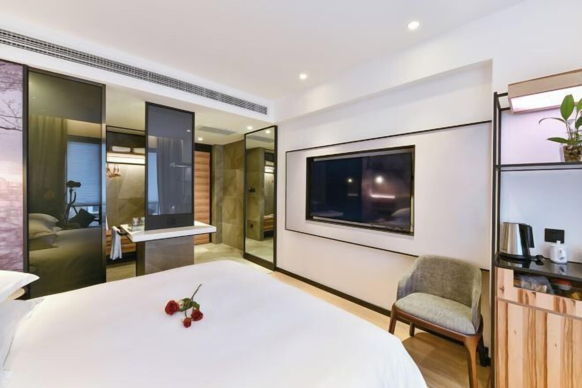 A modern hotel room with a large bed, a bedside table, a chair, a mirror, and a TV.