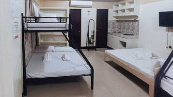 A dormitory room with bunk beds, a kitchenette, a TV, and a cross on the door.