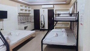 A dormitory room with bunk beds, a TV, a kitchenette, and a cross on the door.
