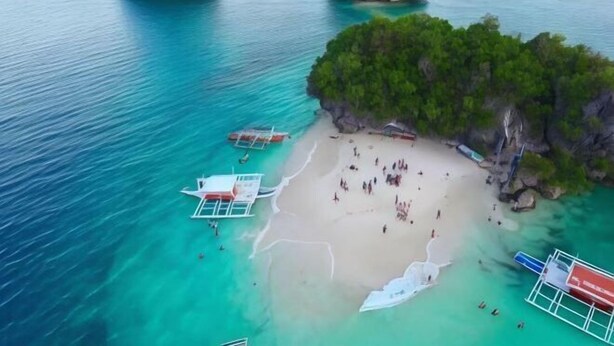 An aerial view of a beach with clear turquoise waters, a sandy shore, and several boats anchored nearby.
