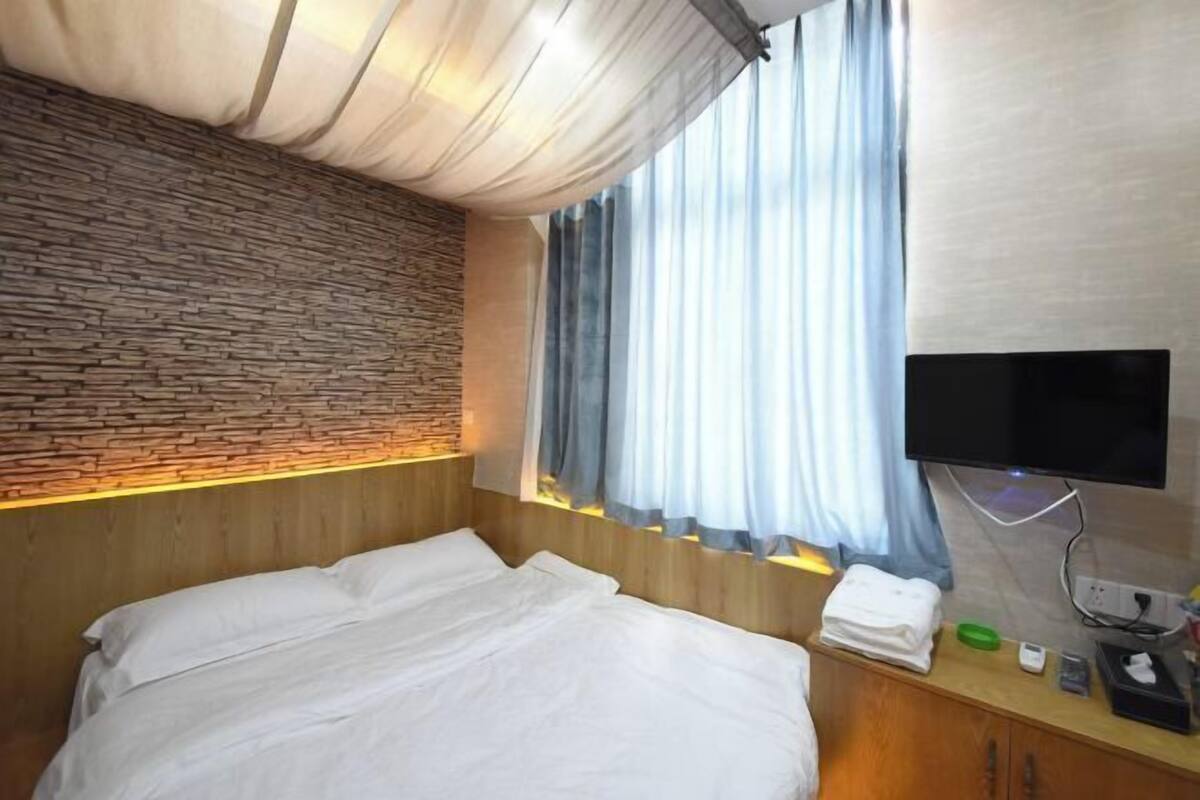 A hotel room with a bed, a flat-screen TV mounted on the wall, a wooden headboard, a stone-patterned wall, and a window with curtains.