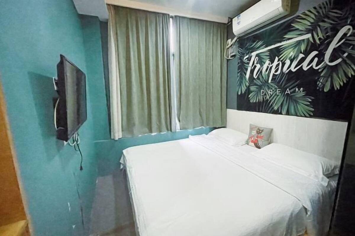 A bedroom with a bed, a TV mounted on the wall, a window with curtains, and a tropical-themed wall decoration.