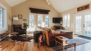 Ein geräumiges Wohnzimmer mit hoher Decke, großen Fenstern, einem Kamin, einem Fernseher und einer gemütlichen Sitzecke.
