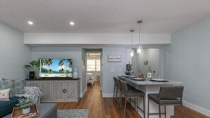 Une cuisine moderne avec un îlot central, un coin repas avec des tabourets de bar, un salon avec un canapé et un téléviseur, et un couloir menant à une chambre.
