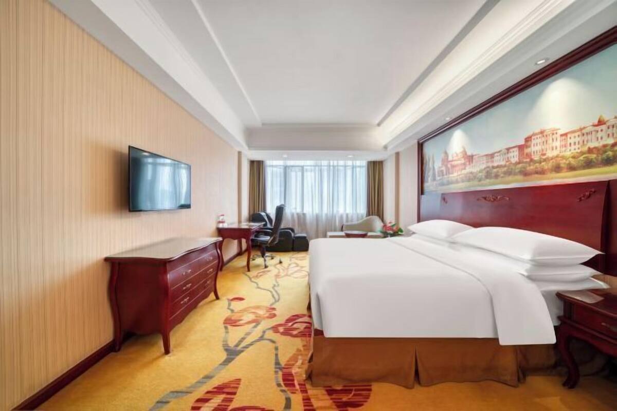 A hotel room with a bed, a desk, a chair, a television, and a painting on the wall.