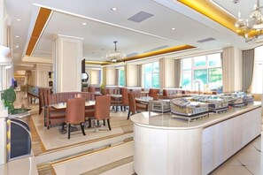 A spacious hotel dining area with multiple seating arrangements, a central bar, and large windows providing natural light.