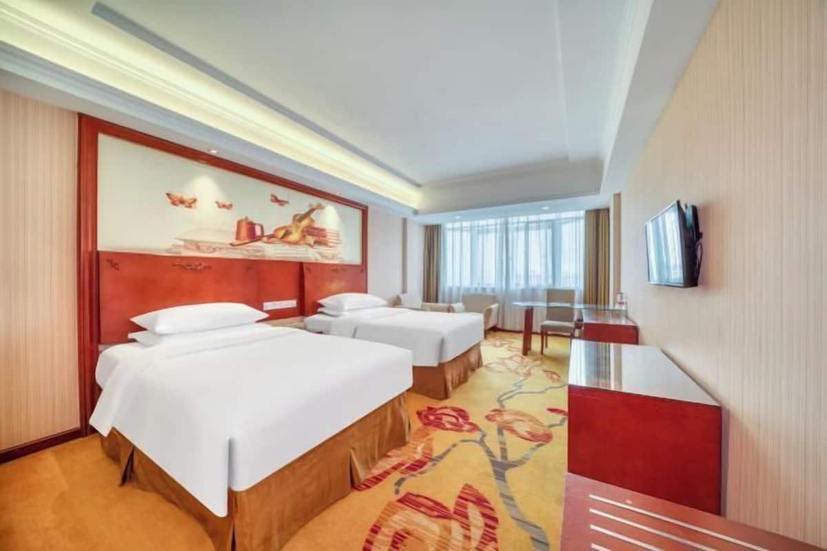 A hotel room with two beds, a desk, a TV, and a large window.