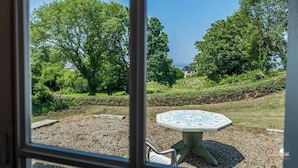 Blick durch ein Fenster auf einen Garten mit einem runden Tisch und Stühlen, umgeben von Bäumen und einer Rasenfläche.