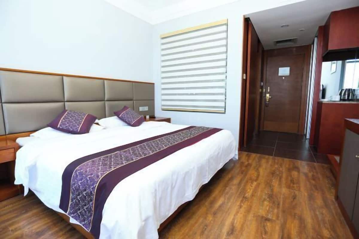 A hotel room with a large bed, a wooden floor, a headboard, a window with blinds, and a door leading to a kitchenette.