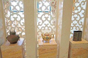 Una habitación con un biombo decorativo que presenta intrincados diseños. Hay dos pequeñas mesas, cada una con un adorno: una maceta de cerámica y un jarrón pequeño.