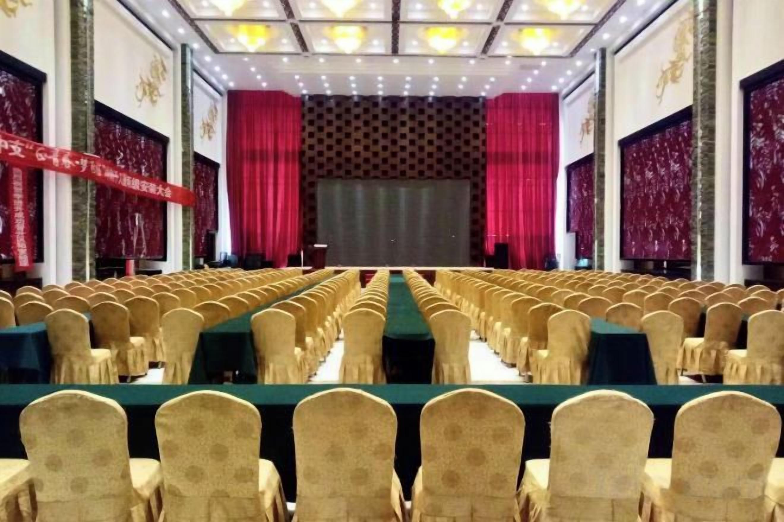Une salle de banquet avec des rangées de tables et de chaises, une scène avec des rideaux et un mur orné de motifs.