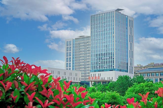 A modern high-rise building with a glass facade, surrounded by greenery and under a partly cloudy sky.