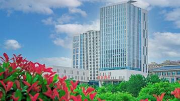 A modern high-rise building with a glass facade, surrounded by greenery and under a partly cloudy sky.