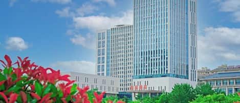 A modern high-rise building with a glass facade, surrounded by greenery and under a partly cloudy sky.