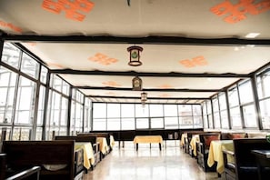 Ein Raum mit Tischen und Bänken, die zum Essen angeordnet sind, großen Fenstern und einer Decke mit einer Hängeleuchte.