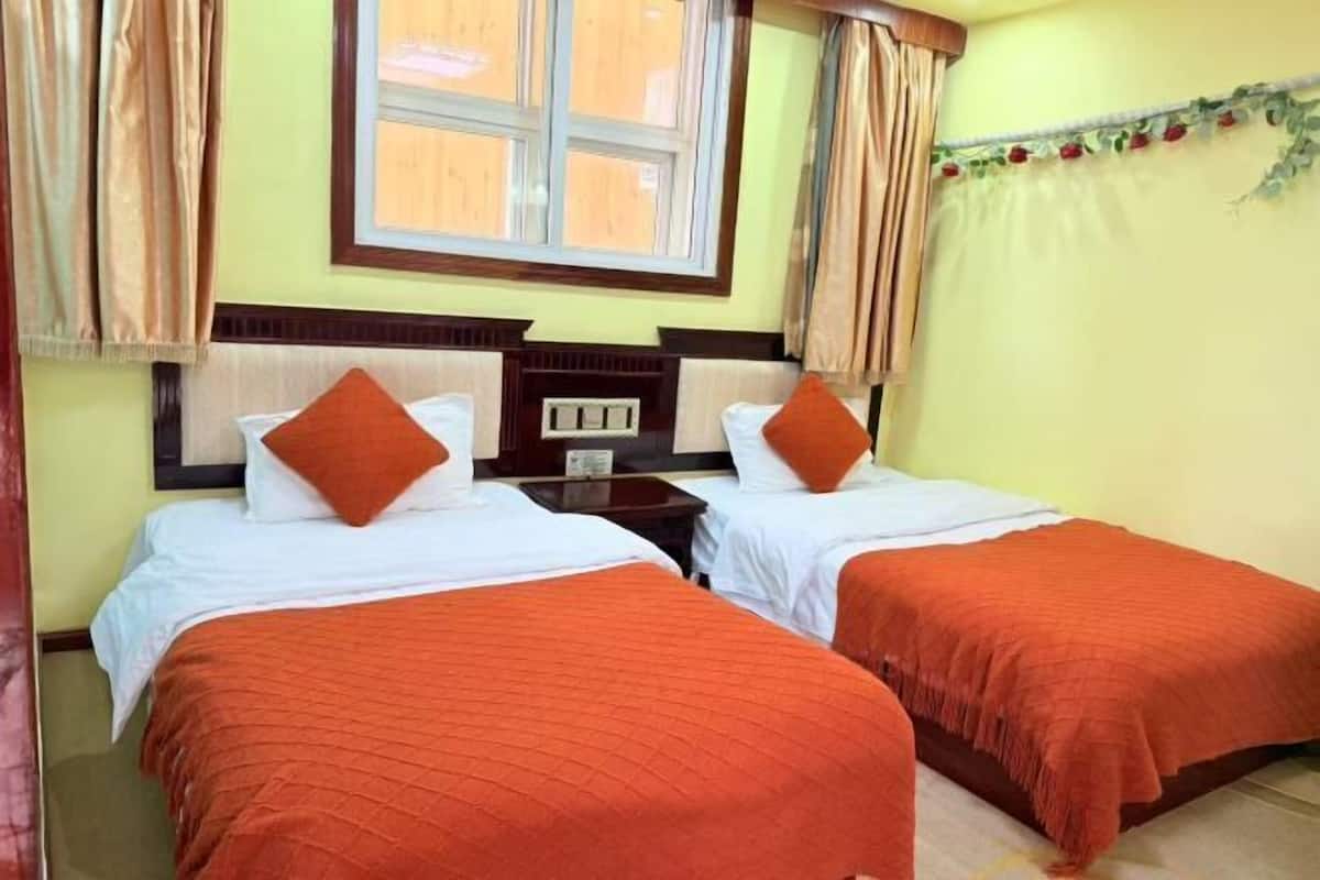 Ein Hotelzimmer mit zwei Betten, jeweils mit orangefarbener Bettdecke und weißen Kissen. Es gibt ein Fenster mit Vorhängen, einen kleinen Tisch mit einer Lampe und eine an der Wand befestigte Uhr.
