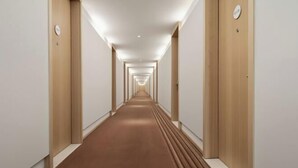 Un pasillo largo y estrecho con puertas de madera a ambos lados, suelo alfombrado y una serie de luces en el techo.