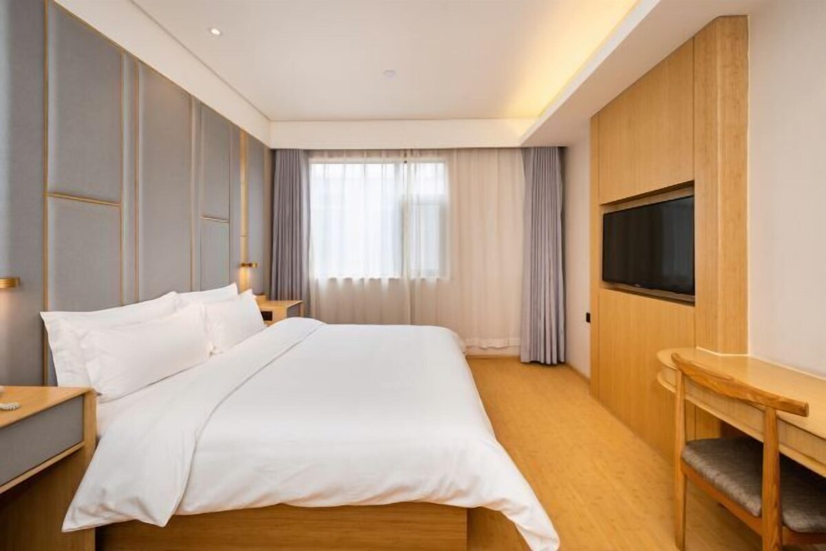 A hotel room with a large bed, wooden headboard, a TV mounted on the wall, and a window with curtains.
