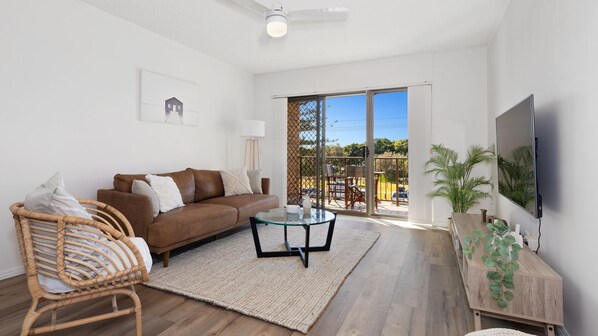 A modern living room with a brown leather sofa, a glass coffee table, a wicker chair, and a TV mounted on the wall.
