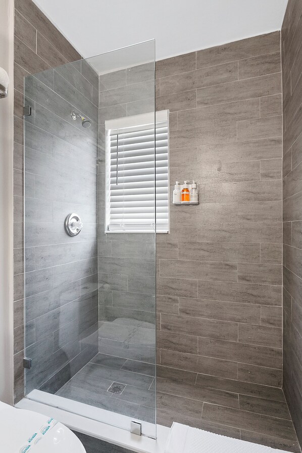 A modern bathroom with a glass-enclosed shower, grey tiled walls, a window with blinds, and a small shelf with toiletries. - ULTRA Modern Apt by the Beach (Hollywood)
