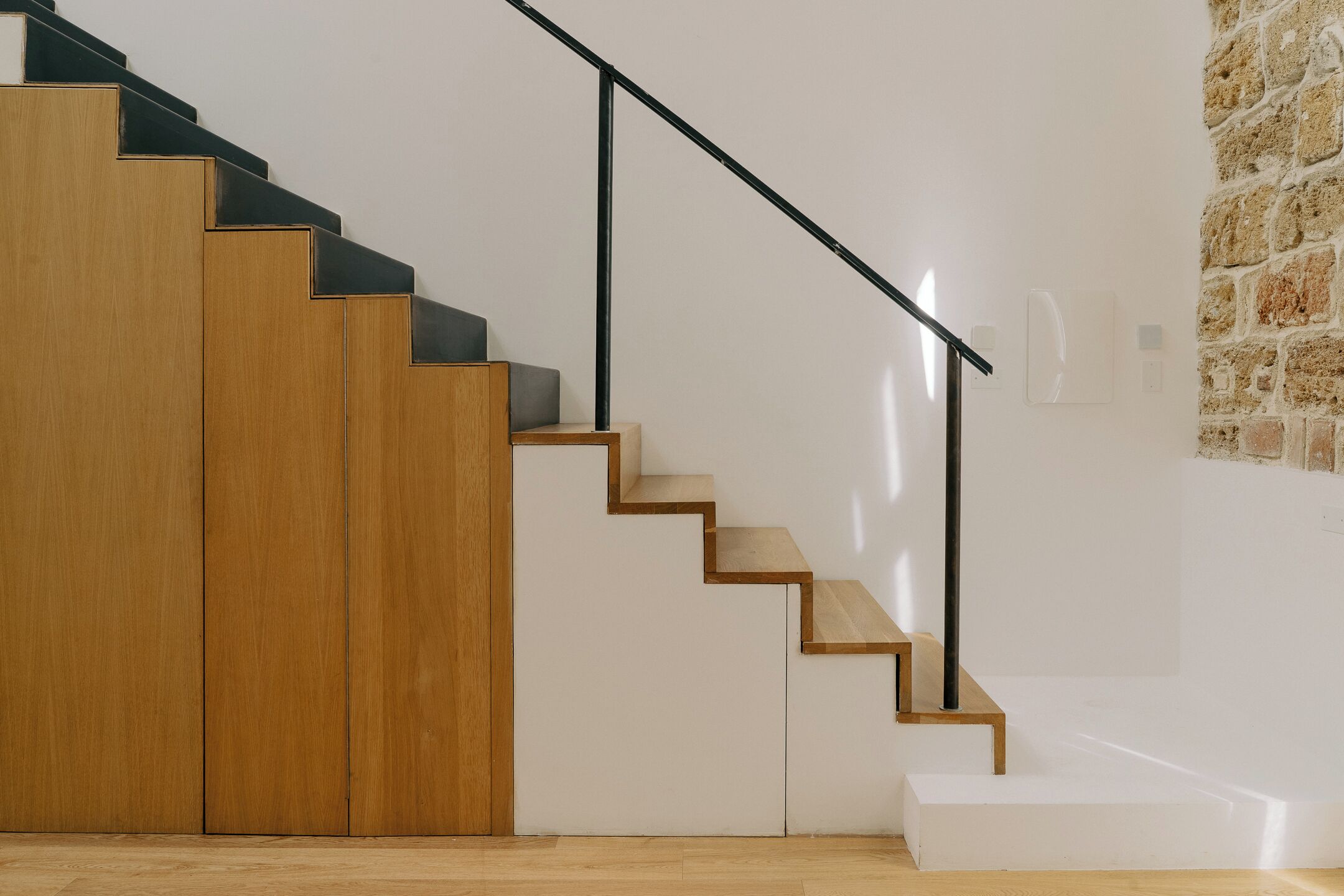 Un escalier avec des marches en bois et des rampes en métal noir, menant à un palier avec un mur blanc et une section de mur en pierre.