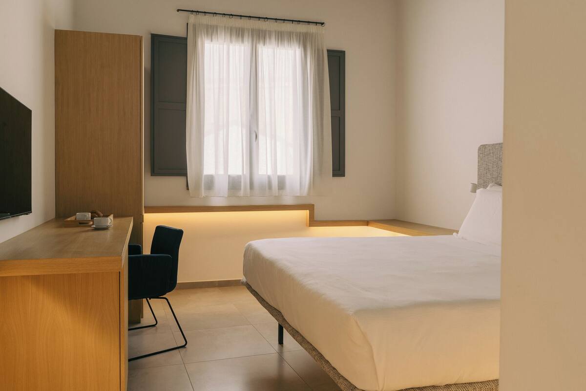 Ein Schlafzimmer mit Bett, Schreibtisch, Stuhl, einem Fenster mit Vorhängen und einem an der Wand befestigten Fernseher.