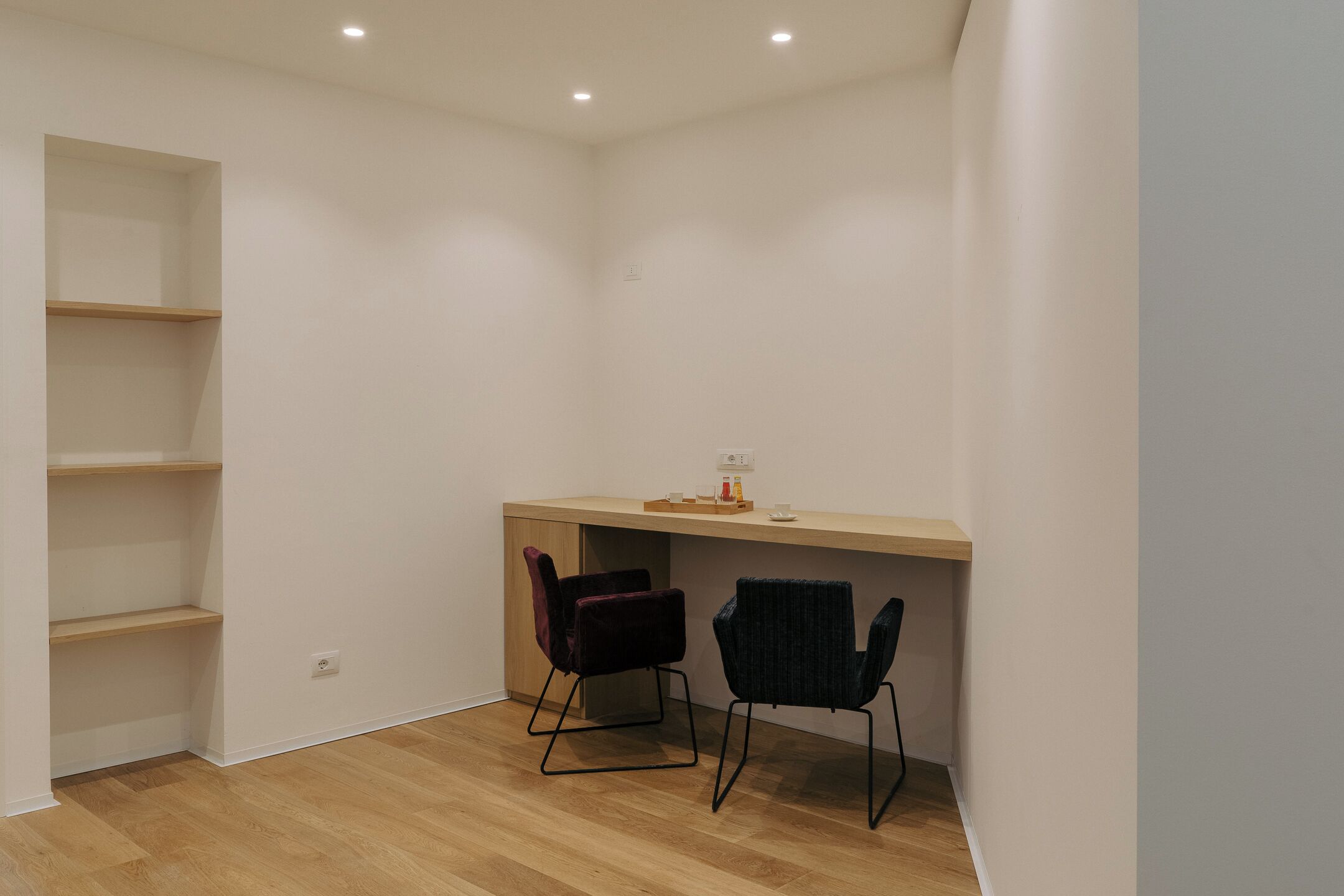 A small, modern room with a wooden desk and two chairs, a built-in shelf, and a light switch on the wall.