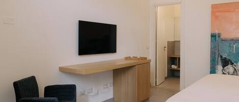 A modern hotel room with a bed, a desk with a flat-screen TV, two chairs, and a painting on the wall.