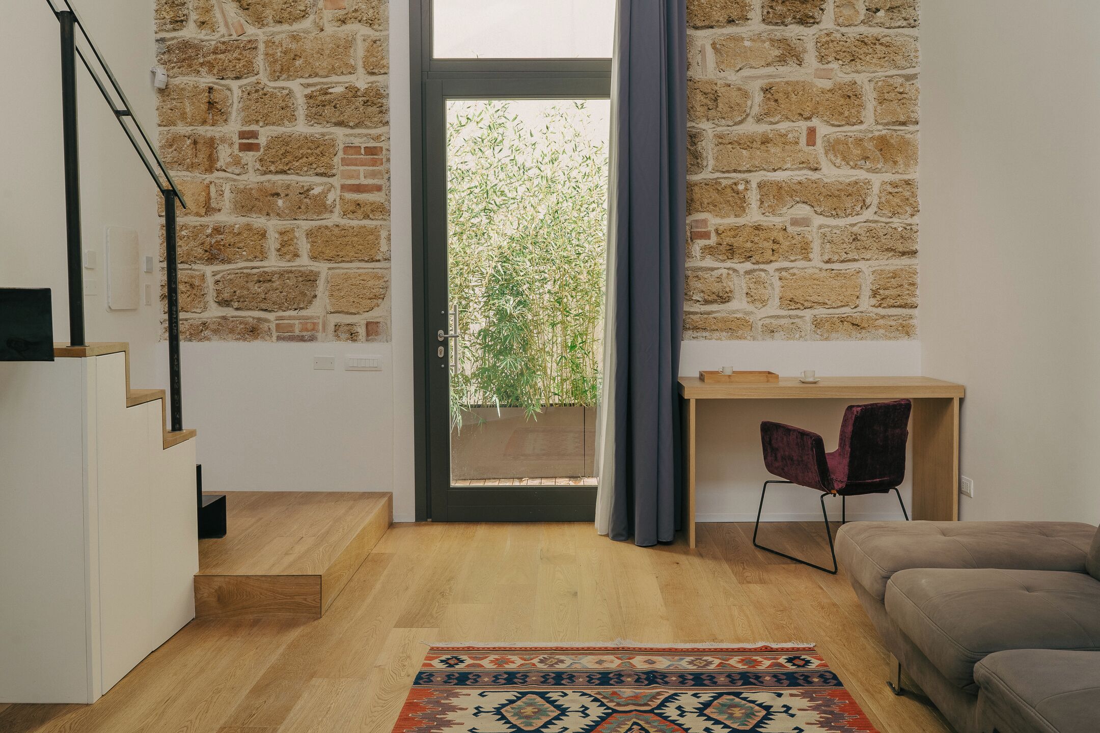 Un salón moderno con sofá, un escritorio de madera y una escalera que lleva a un balcón con vistas a la vegetación.