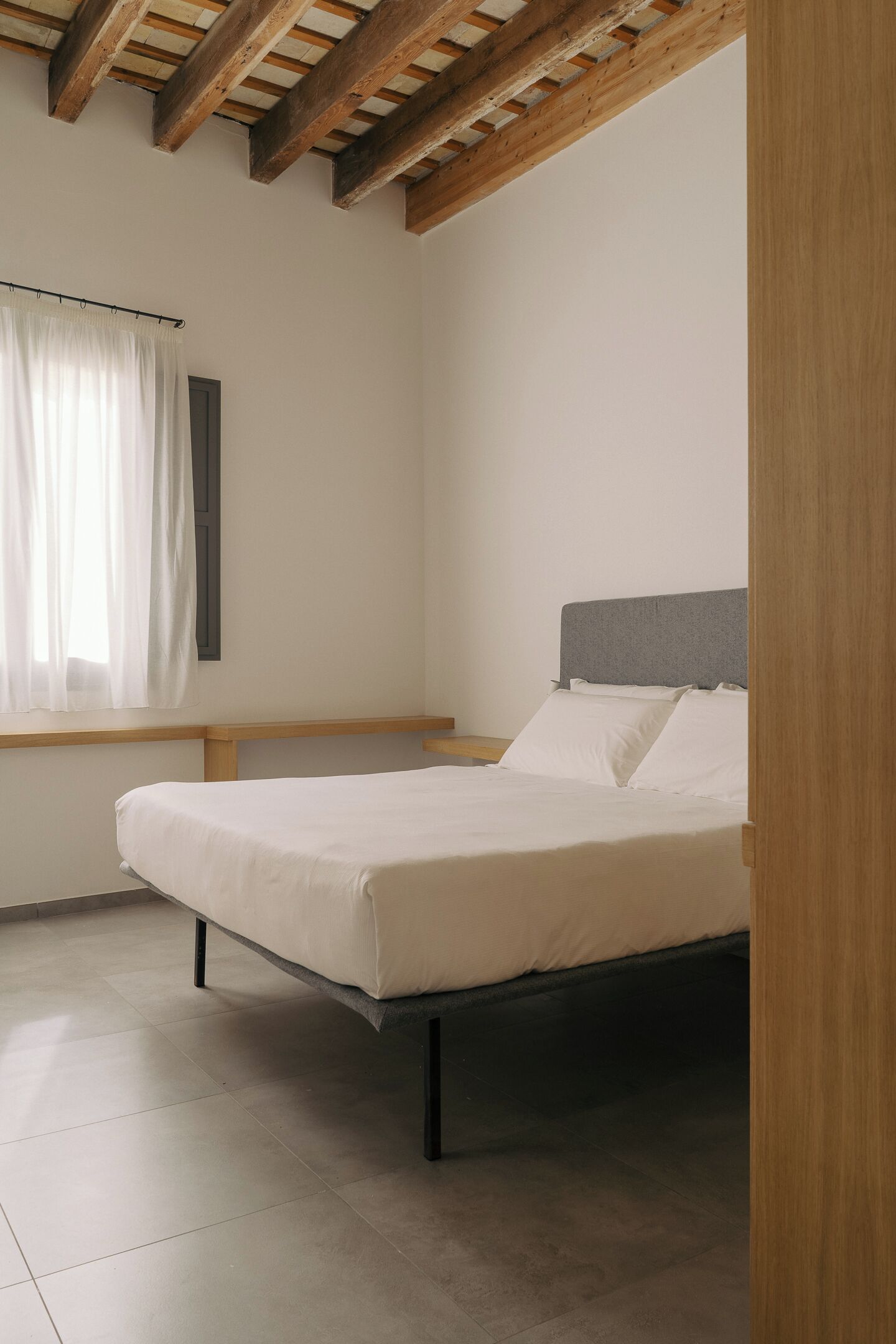 A bedroom with a bed, wooden beams, a window with curtains, and a plain white wall.