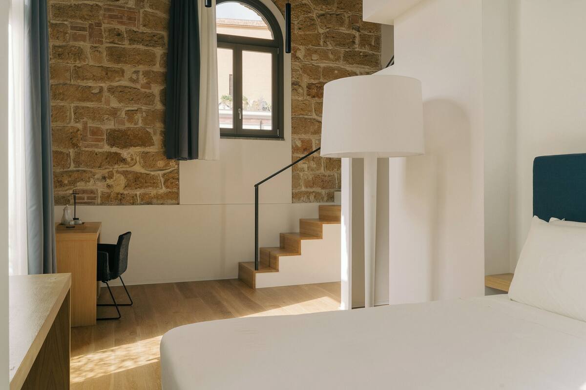 Ein Schlafzimmer mit einem großen Bett, einem Schreibtisch mit Stuhl, einer Treppe und einem Fenster mit Vorhängen.