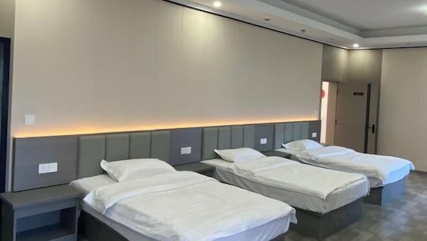 Habitación de hotel con tres camas individuales, una mesita de noche y una lámpara de pared.