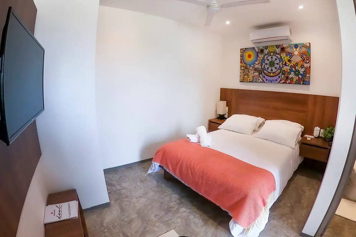 A hotel room with a bed, a nightstand, a TV, an air conditioning unit, and a ceiling fan.