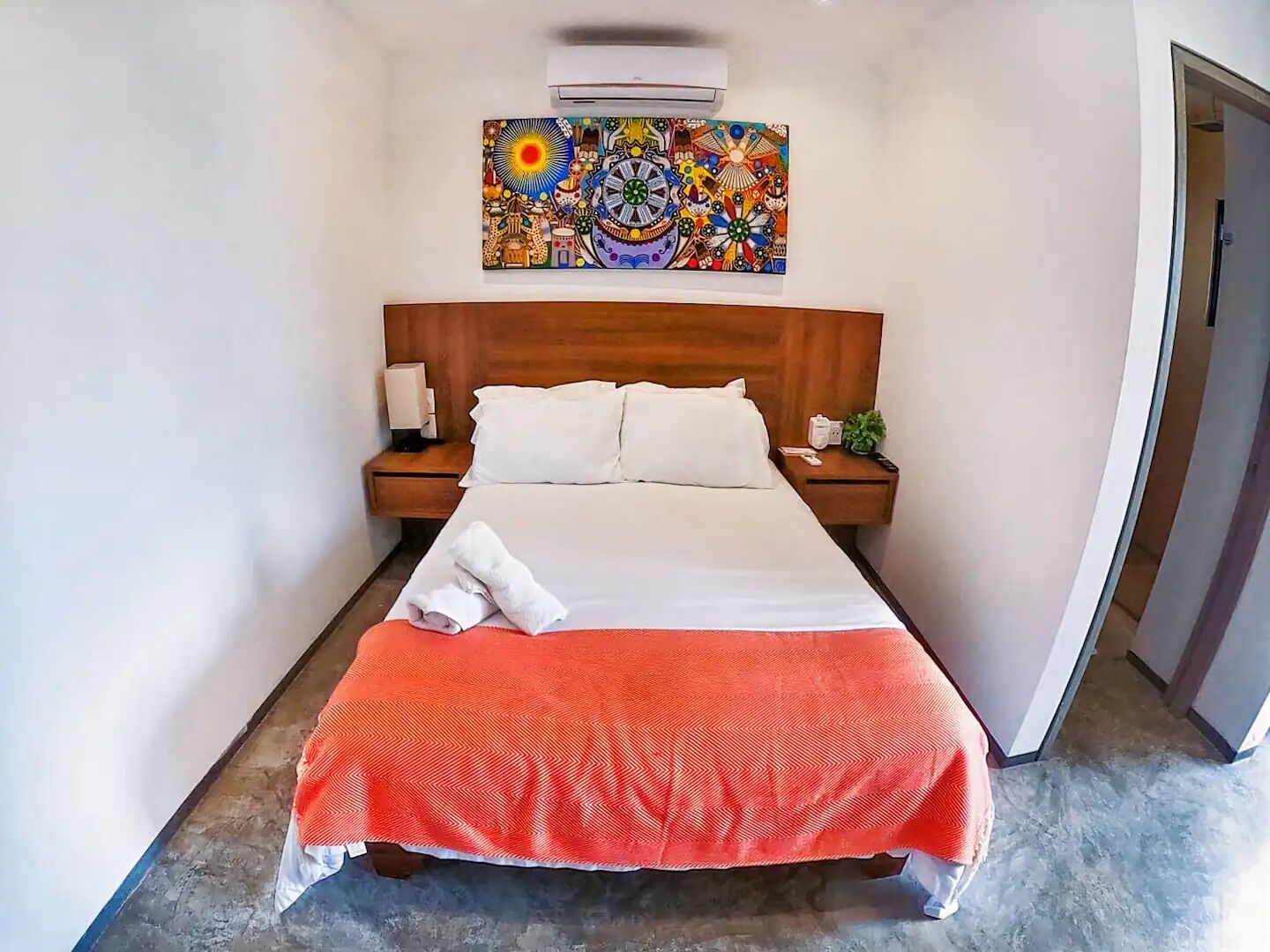 A hotel room with a bed, two bedside tables, a nightstand, a wall-mounted air conditioner, and a colorful painting above the bed.