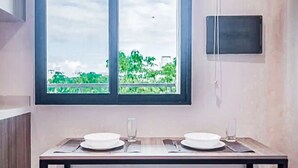 A small dining area with a table and two chairs, a window with a view of greenery, and a wall-mounted TV. - El Mexicano (Playa del Carmen)