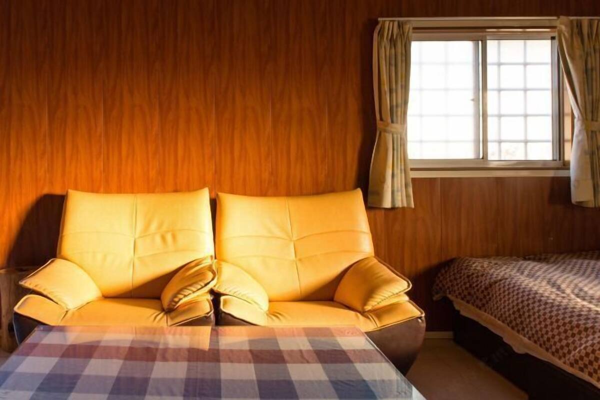 A room with a wooden wall, a sofa with cushions, a checkered tablecloth, and a bed with a patterned blanket.