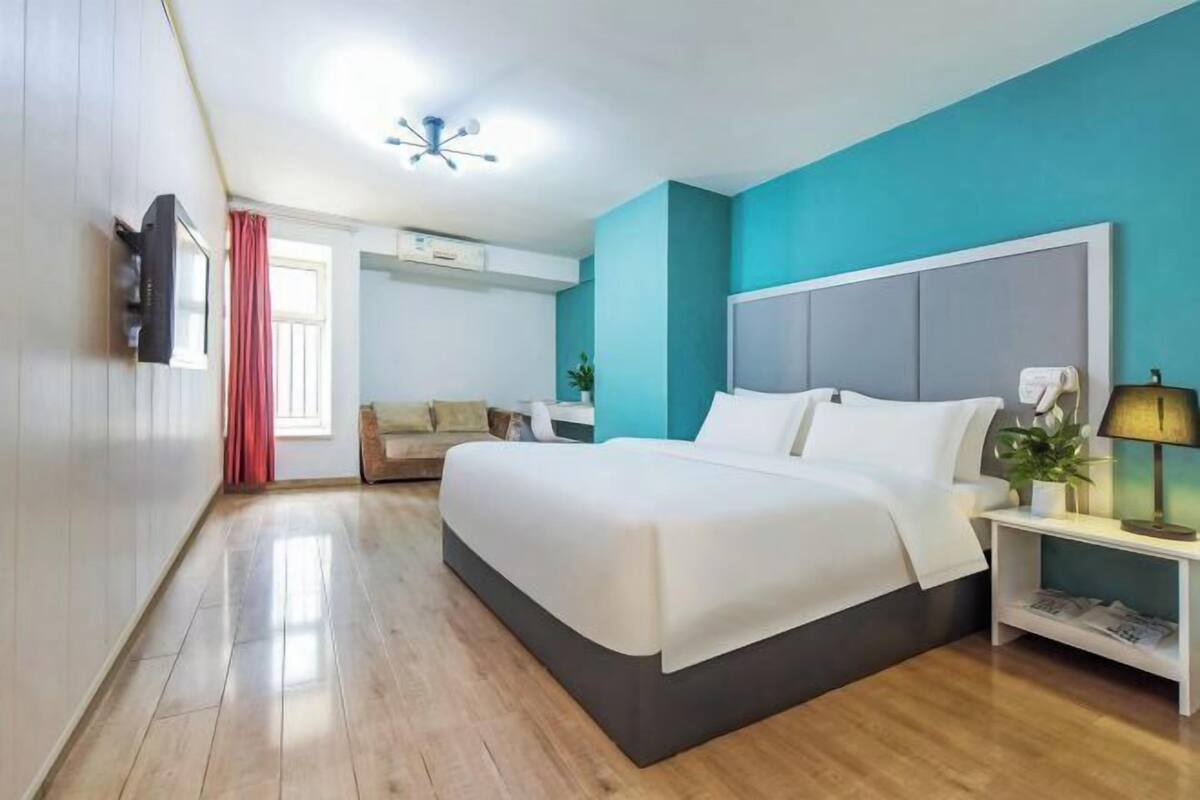 A modern hotel room with a large bed, a TV, a bedside table with a lamp, and a window with curtains.
