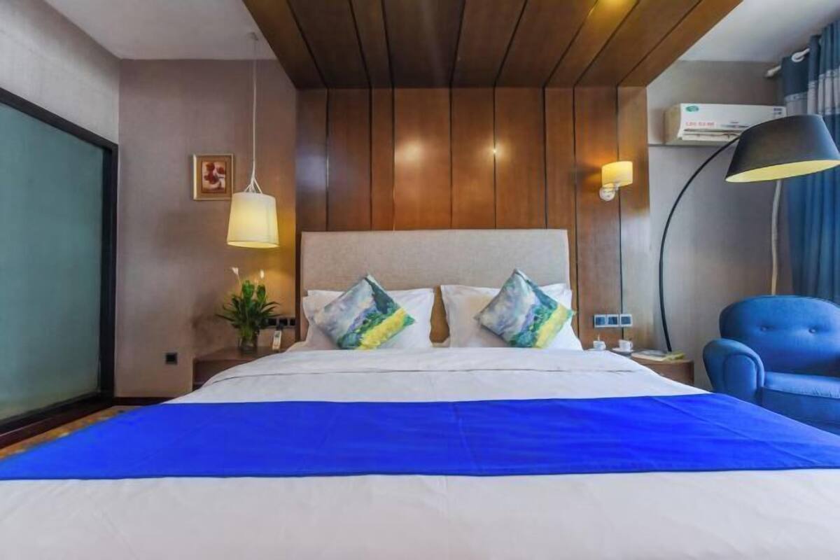 A hotel room with a large bed, a blue bedspread, a wooden ceiling, a wall-mounted air conditioner, a blue armchair, and a bedside table with a lamp.