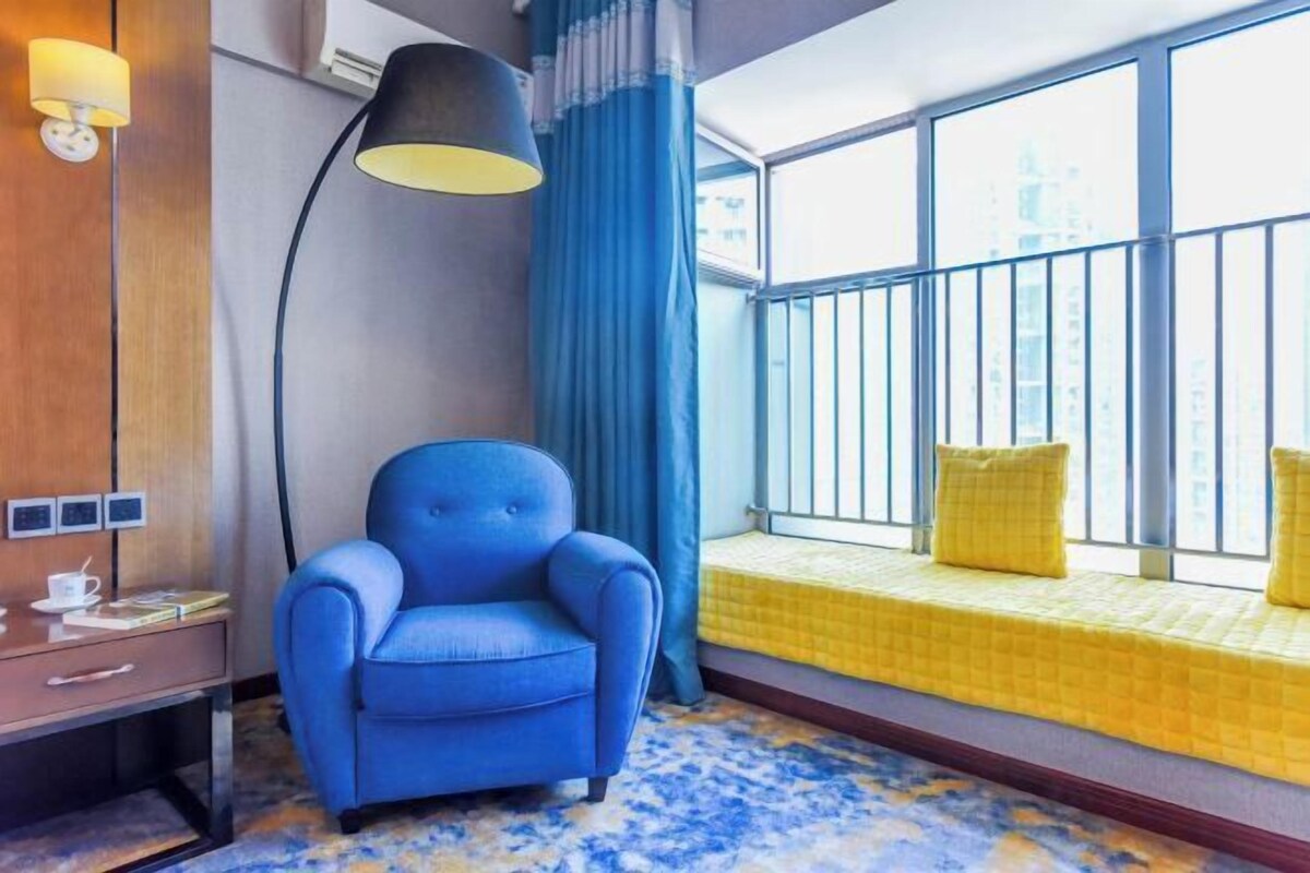 A hotel room with a blue armchair, a small desk with a lamp, a window with a yellow bed, and a city view.