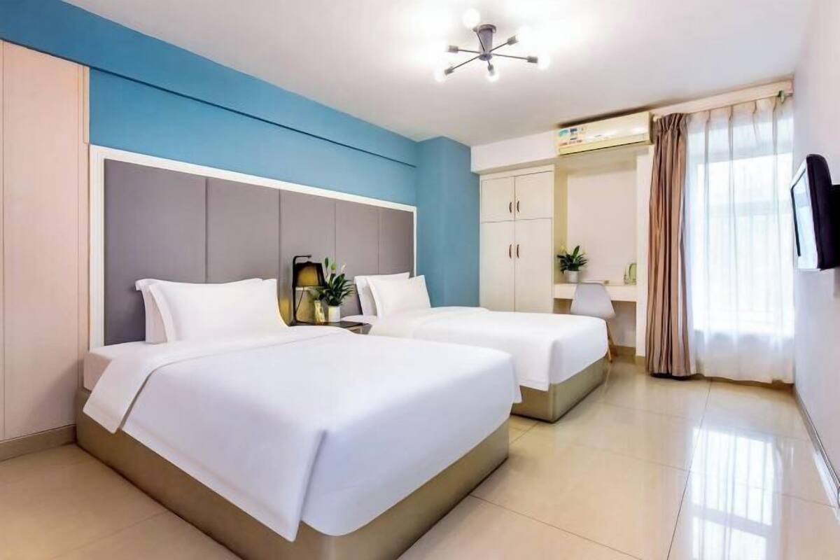 A hotel room with two beds, a ceiling fan, a wardrobe, a desk with a lamp, and a television.